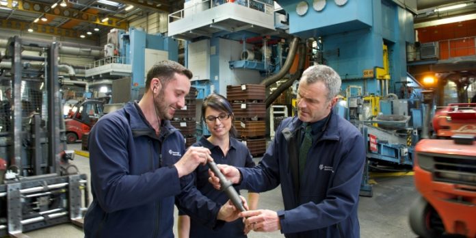 Aufmacher © Hirschvogel Automotive Group Produktion von Hirschvogel: Werkleiter und Mitarbeiter begutachten eine mehrstufig automatisch kalt umgeformete Getriebewelle. Im Hintegrund stehen mehrere Pressenaggregate.