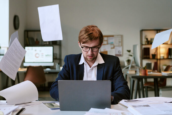 Young Caucasian Man Working at Desk Surrounded by Flying Papers in Office